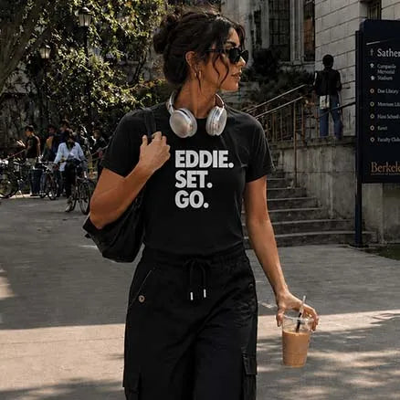 A woman walking outdoors on a campus-style path wearing a black graphic T-shirt with the text 'EDDIE. SET.