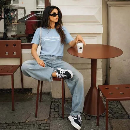 A woman sits on a brown metal stool at an outdoor cafe table, wearing a light blue graphic t-shirt, light-wash wide-leg jeans, black and white sneakers, and sunglasses.