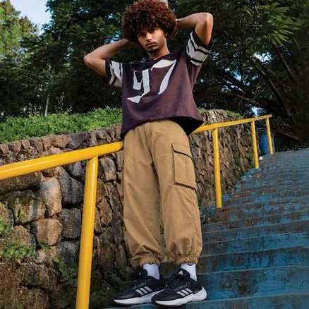 A person standing on outdoor blue stairs next to a stone wall and yellow railing, wearing a dark oversized graphic T-shirt, tan cargo pants, white socks, and black-and-white sneakers.