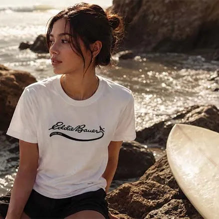 A woman sitting on a rocky beach wearing a white t-shirt with a black 'Eddie Bauer' logo, with ocean waves and a surfboard in the background.