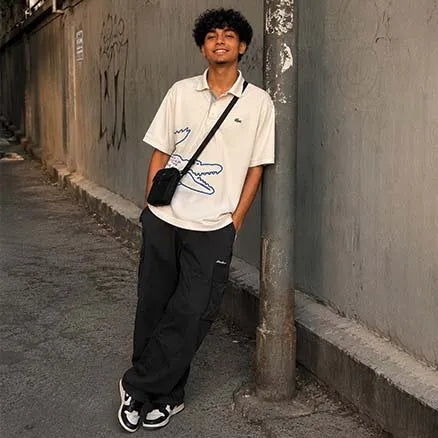 A man wearing a cream Lacoste polo shirt with a blue crocodile graphic, black cargo pants, and a black crossbody bag, leaning against a concrete wall in an alley.