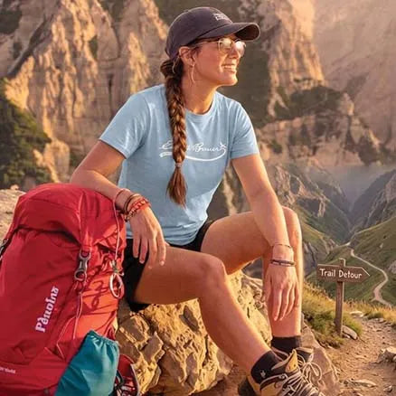 A woman hiking in a mountain setting, wearing a light blue crewneck t-shirt with a white script logo, black athletic shorts, and a dark baseball cap. She is sitting on a rock next to a red Patagonia hiking backpack, with a trail sign reading 'Trail Debour' in the background.
