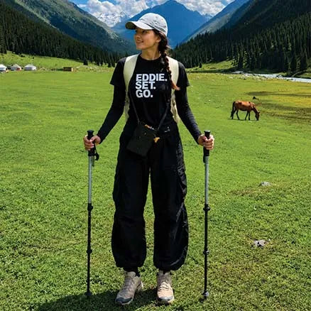 A woman stands in a mountain meadow holding hiking poles, wearing a black graphic T-shirt with 'EDDIE. SET.