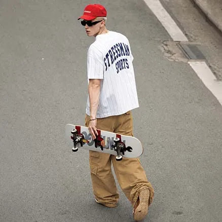 A person walking on an asphalt street holding a skateboard under their arm. They are wearing a white pinstripe short-sleeve t-shirt with 'STRECHING SPORTS' printed in navy blue on the back, tan cargo pants, a red baseball cap, and sunglasses.