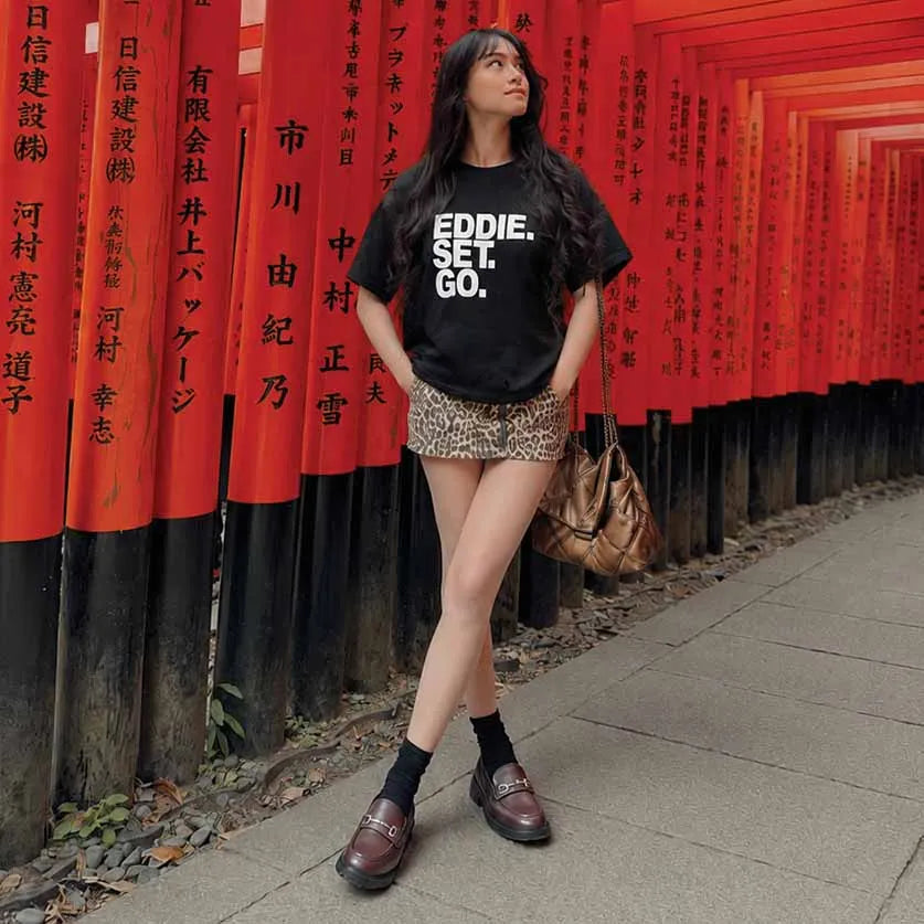 A woman stands in front of a row of red torii gates wearing a black graphic t-shirt with 'EDDIE. SET.