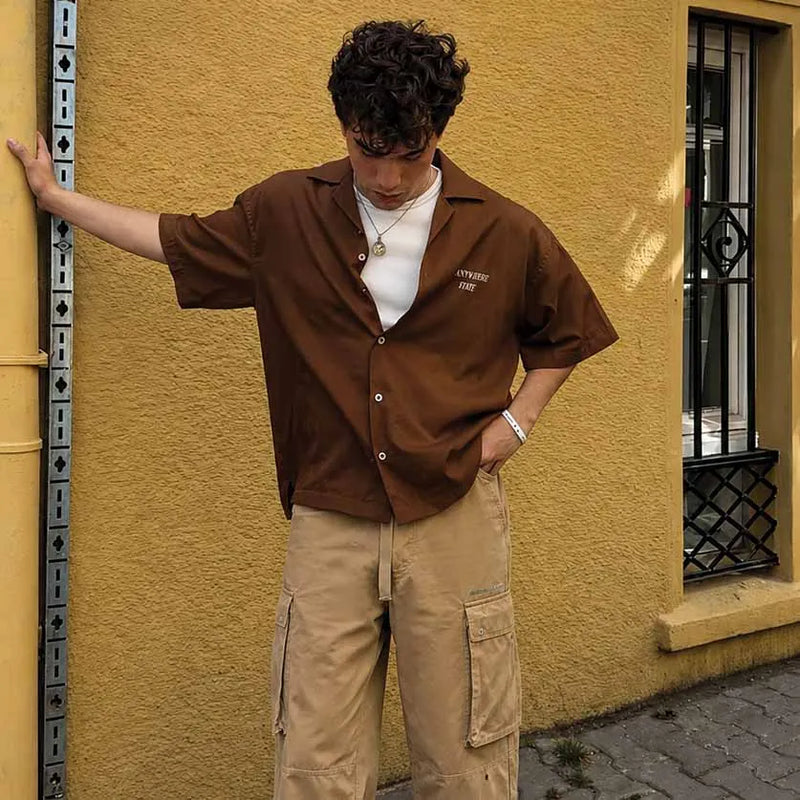 A man wearing a brown short-sleeve button-up shirt over a white t-shirt and beige cargo pants, leaning against a textured yellow wall.