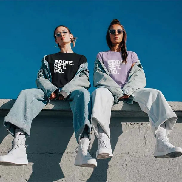 Two women sitting on a concrete ledge against a clear blue sky, wearing oversized light-wash denim sweatpants and sweatshirts with 'EDDIE SET GO.' printed in white.
