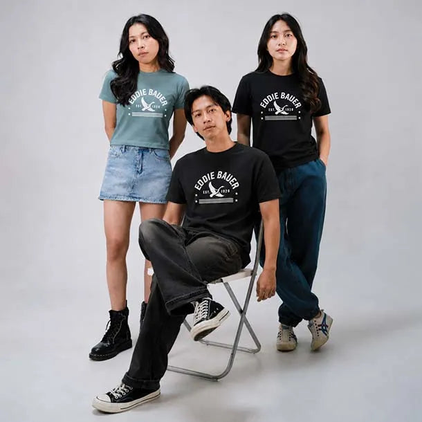 Three models posing in a studio setting wearing Eddie Bauer t-shirts. The seated model wears a black crew-neck t-shirt, while the standing models wear one in sage green and one in black.