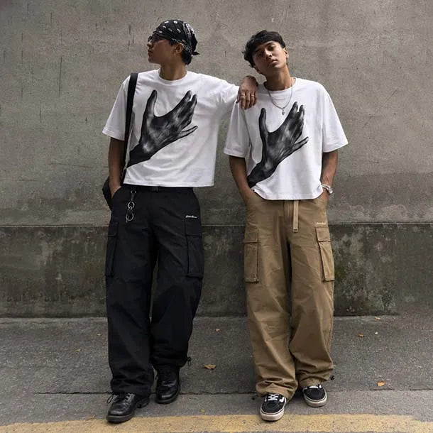 Two men standing outdoors against a concrete wall wearing matching white graphic T-shirts with a large black hand print on the front, styled with different cargo pants and accessories.